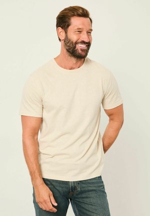 Smilende mand med brunt hår og skæg iført beige t-shirt og blå jeans, stående mod en ensfarvet lys baggrund.