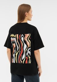 Černé oversized tričko s barevným abstraktním potiskem na zadní straně, s vlnitými tvary v červené, žluté, bílé a černé barvě.