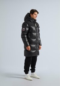 Muž v dlhom čiernom puffer kabáte s kapucňou, čiernych teplákach a bielych teniskách, stojí proti jednému svetlému šedému pozadiu.