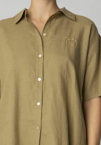 Khakifarbene Leinenbluse mit Knopfverschluss und kurzen Ärmeln. Verfügt über ein dezentes gesticktes Logo auf der Brust und weiße Knöpfe.