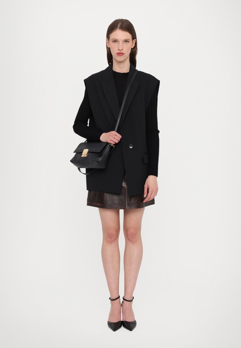 Sort oversized vest med revers over en sort bluse med lange ærmer, parret med en brun læder miniskirt og sorte stiletto hæle, mens hun holder en sort taske.