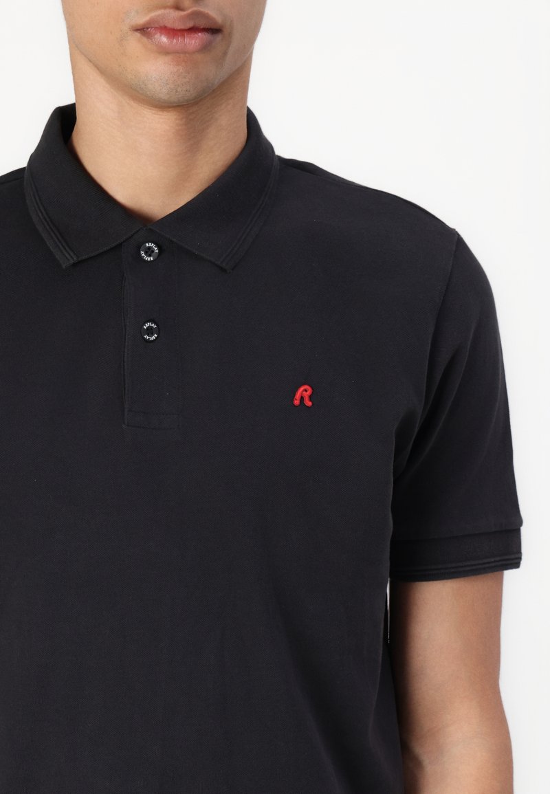 Polo negro de algodón con cuello, cuenta con dos botones, mangas cortas y un pequeño logotipo rojo "R" en la parte izquierda del pecho.