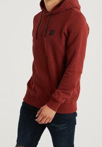 Sudadera roja hecha de tela suave, con un bolsillo frontal, capucha con cordón y un pequeño emblema negro en el pecho. Lleva puestos jeans oscuros.