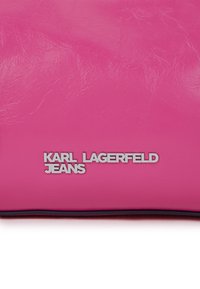Φωτεινή ροζ δερμάτινη επιφάνεια με μεταλλικό λογότυπο "KARL LAGERFELD JEANS" σε ασημί χρώμα, τυπωμένο κοντά στο κάτω κέντρο.