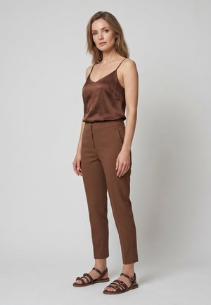 Vrouw staand, gekleed in een bruine satijnen hemd, bijpassende bruine smalle broek en bruine sandalen met bandjes tegen een effen grijze achtergrond.