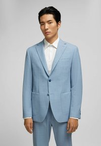 Lichtblauwe colbert gemaakt van textiel met textuur, voorzien van twee voorkussens, een notch revers en een enkele knoopsluiting. Draagt over een wit shirt.