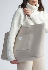 Bolsa de tela beige con un gran bolsillo exterior, asas de textil y una etiqueta. El modelo lleva un suéter blanco de canalé y pantalones claros.