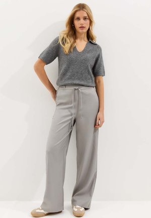 Kobieta ubrana w szary krótki sweter z dzianiny, jasnoszare spodnie o szerokich nogawkach z wysokim stanem i wiązaniem oraz beżowo-białe trampki stoi na tle białej ściany.