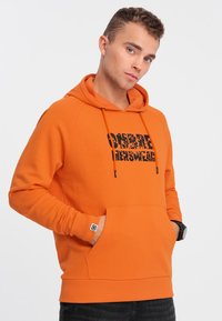 Felpa arancione con cappuccio e testo grafico nero "OMBRE" sul davanti, tasca a marsupio e polsini a costine. Materiale morbido, vestibilità rilassata.