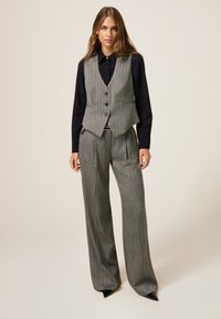 Grijze vest met patroon en knopen, gecombineerd met bijpassende grijze wijde broek. Zwarte blouse eronder. Gladde stof met op maat gemaakt ontwerp.