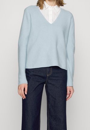 Persoon draagt een lichtblauwe V-hals trui over een wit overhemd met kraag, gecombineerd met donkere blauw wijde broekspijpen jeans.