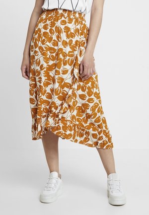 Femme portant une jupe portefeuille fleurie blanche et orange associée à des baskets blanches, debout devant un fond clair uni.