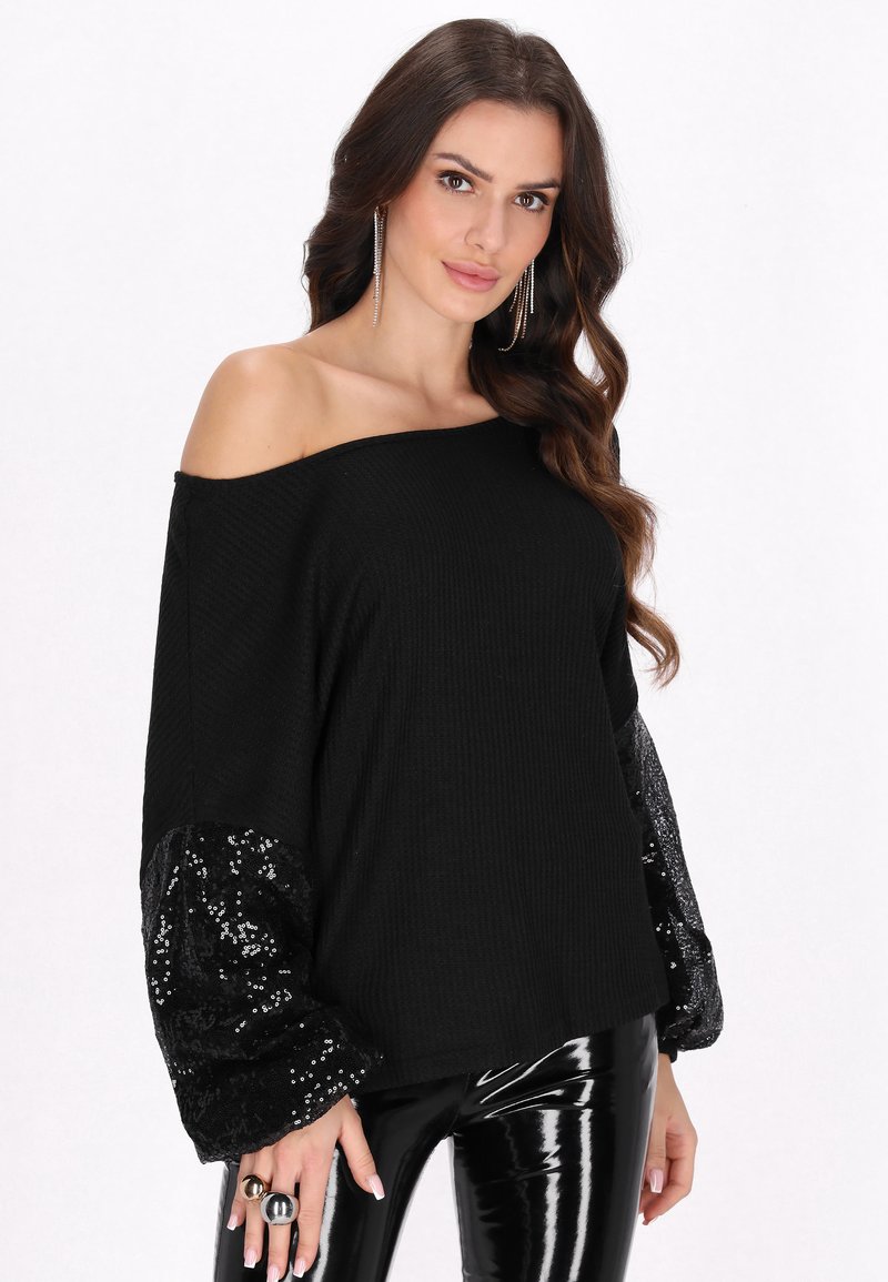 Maglione nero off-shoulder con texture a coste e maniche a palloncino in paillettes. Abbinato a pantaloni attillati neri lucidi.