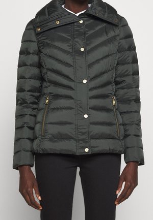 Frau trägt schwarze gesteppte Jacke mit goldfarbenen Druckknöpfen und Reißverschlusstaschen, steht vor einem schlichten Hintergrund.