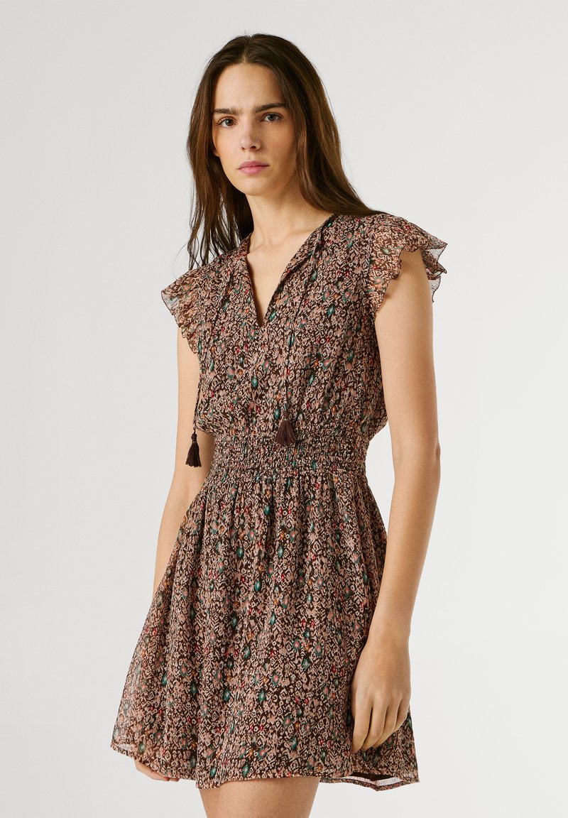 Pikkade pruunide juustega naine, kes kannab lühikeste varrukatega mustrilist kleiti, millel on liibuv vöökoht ja kaelaesisele kinnitatavad narmastega nöörid, seisab ühevärvilise tausta ees.