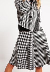 Ensemble ve vzoru černobílé houndstooth skládající se z dobře padnoucí bundy s velkými knoflíky a rozšířenou sukní, vyrobené z texturované látky.