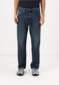 Jean en denim bleu foncé avec une coupe droite, cinq poches et une fermeture à bouton. Porté avec des chaussures de sport grises.