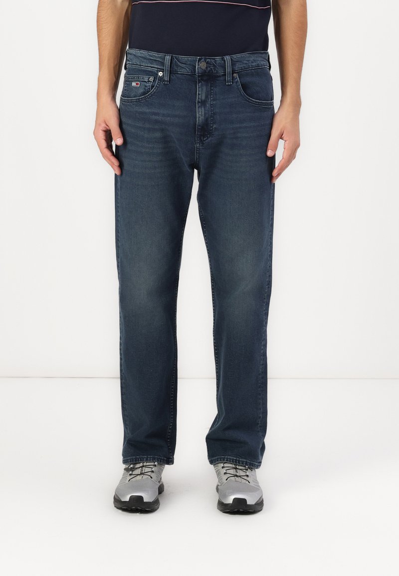 Jean en denim bleu foncé avec une coupe droite, cinq poches et une fermeture à bouton. Porté avec des chaussures de sport grises.