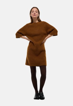Robe en tricot marron avec une silhouette décontractée, des manches courtes larges et un ourlet au-dessus du genou. Le modèle porte des mocassins noirs et des collants.