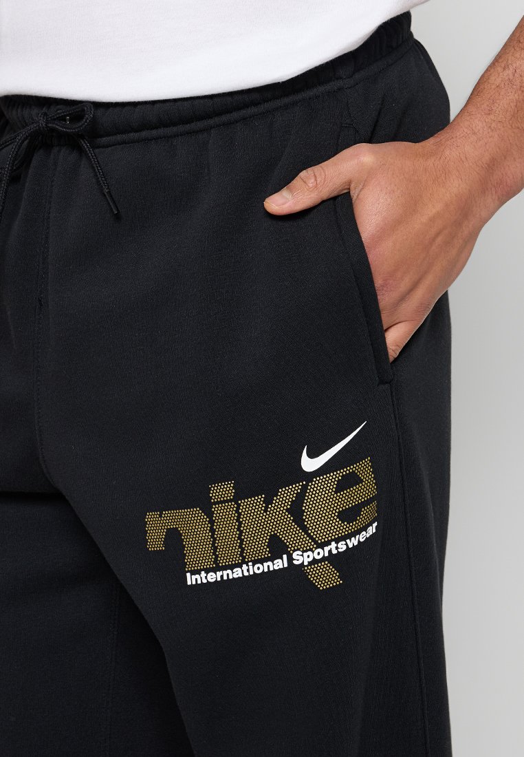Main dans la poche d'un pantalon de survêtement noir Nike avec le texte "nike" en or en pointillés et les logos "International Sportswear" et swoosh en blanc.