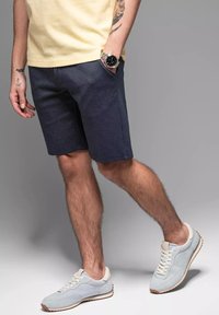 Pantaloncini blu scuro, vestibilità regolare, realizzati in tessuto simile al denim. Abbinati a sneakers grigie chiare e una maglietta gialla. Orologio al polso sinistro.