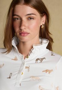 Biele košele s golierom, na ktorej sú vzory zvieracích a čižmových odtlačkov v hnedých odtieňoch, s volánovým golierom a zlatými gombíkmi na zapínanie.