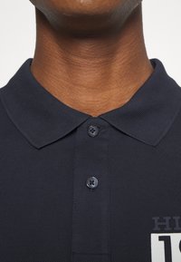 Marine polo met een gestructureerde stof, twee zwarte knopen op de kraag en een witte bedrukte accent op de borst.