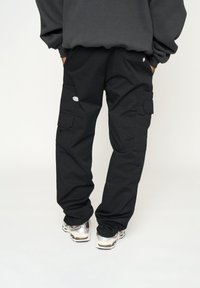 Pantalones cargo negros con múltiples bolsillos, corte holgado y puños elásticos. Fabricados con tejido resistente. Detalle de logo plateado en el bolsillo.