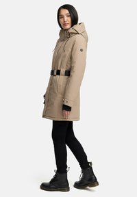 Manteau isolé beige avec capuche, ceinture noire, poches latérales et poignets à boutons, porté sur des leggings noirs et des bottes noires à lacets.