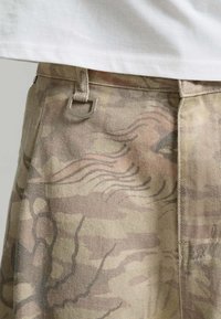 Gros plan sur un pantalon beige avec des motifs abstraits de visage et de vague, avec des passants de ceinture et un anneau métallique argenté en forme de D.