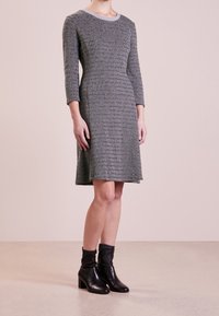 Donna che indossa un vestito grigio a maniche tre quarti, lungo fino al ginocchio, con stivaletti neri con tacco, in piedi su un pavimento chiaro.