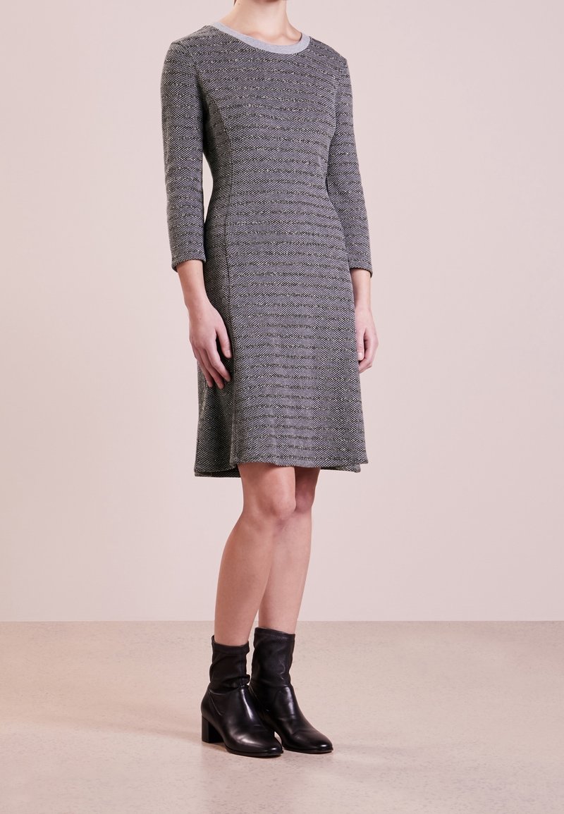 Donna che indossa un vestito grigio a maniche tre quarti, lungo fino al ginocchio, con stivaletti neri con tacco, in piedi su un pavimento chiaro.