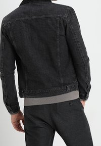 Chaqueta de denim negro con cuello con solapa, costuras visibles y diseño ajustado. Combinada con sudadera gris claro con ribete acanalado en el bajo.
