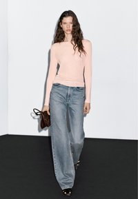 Hellrosa Langarmoberteil kombiniert mit weitem blauen Jeans. Das Modell hält eine braune Tasche. Die Szene ist vor einer schlichten weißen Wand mit einem dunklen Boden eingerichtet.