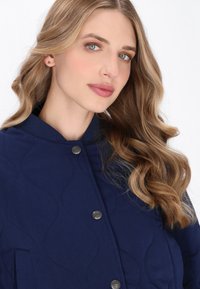 Femeie blondă cu păr ondulat, îmbrăcată într-o jachetă cu model matlasat de culoare navy și nasturi metalici, privind ușor spre lateral pe un fundal alb.