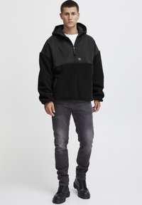 Svart pulloverjacka med en texturerad, fluffig underdel och en slät överdel. Har en huva, halvdragkedja och en logotyp patch.