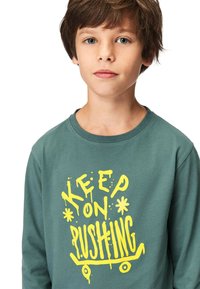 Grünes Langarm-T-Shirt mit gelbem Graffiti-ähnlichem Text "KEEP ON PUSHING" und Skateboard-Grafik. Baumwollstoff, runder Ausschnitt.