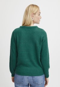 Maglione verde con un motivo a maglia testurizzata, polsini e orlo a costine, e un classico scollo rotondo. Indossato con jeans blu.