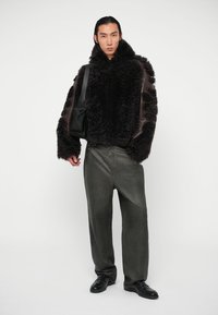 Hombre vistiendo una chaqueta de piel de oveja negra, pantalones oscuros grises holgados, zapatos negros y llevando un bolso de hombro negro, de pie contra un fondo blanco.