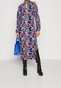 Vestido floral de longitud media en azul marino con flores rojas y rosas, con mangas largas, escote de ojal y aberturas laterales. Combinado con un bolso azul y botas negras.
