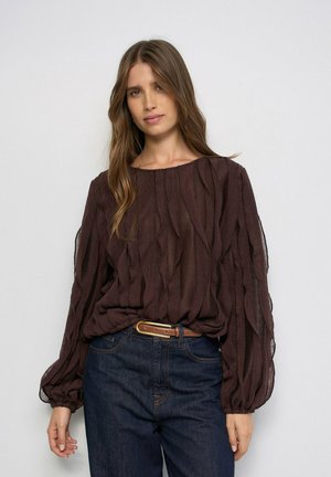 Mujer con cabello largo y castaño que lleva un suéter de punto trenzado marrón oscuro con mangas abullonadas y vaqueros azul oscuro sobre un fondo liso.