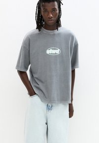 Graues, übergroßes T-Shirt mit rundem Halsausschnitt und Logo auf der Vorderseite. Der Baumwollstoff hat eine glatte Textur und wird mit hellblauen Jeans kombiniert.