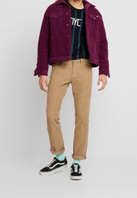 Bordeaux fleecejacka, svart grafisk t-shirt med blå ränder, beige kordflamsbyxor, svarta sneakers med vita sulor, och mintgröna strumpor.