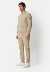 Beige knitted sweater with a round neckline and ribbed cuffs, layered over a checkered shirt, paired with light beige chinos and white sneakers.