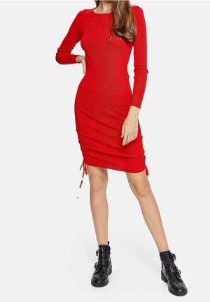 Frau trägt ein figurbetontes, langärmeliges rotes Kleid mit verstellbaren Seitenschnüren und schwarze Stiefeletten mit Schnalle, steht mit einer Hand in der Hüfte.