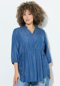 Blusa in denim blu con scollatura a V, maniche 3/4, vita arricciata con lacci e spacchi laterali per facilitare il movimento.