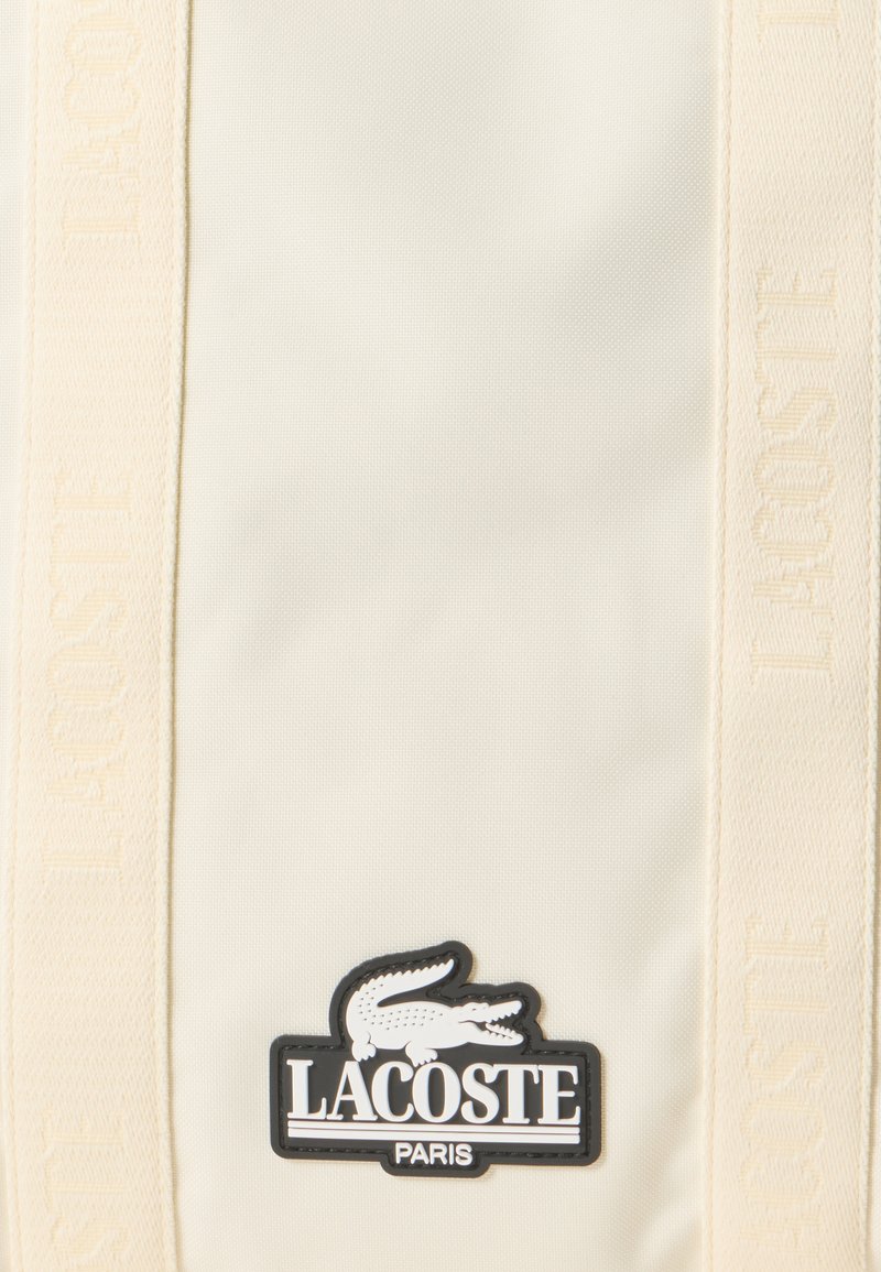 Tessuto beige con strisce verticali tessute e un logo patch nero che mostra un coccodrillo bianco e il testo "LACOSTE PARIS."