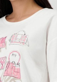 Bílá mikina s růžovými grafickými ilustracemi různých kabelčeků, které mají odlišné tvary a designy, umístěné na prsou.