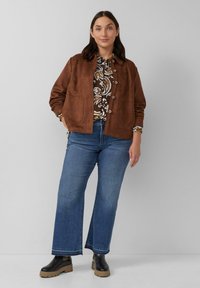 Frau in brauner Wildlederjacke, gemustertem Hemd, blauen weiten Jeans und schwarzen Stiefeln, die vor einem schlichten, hellen Hintergrund steht.