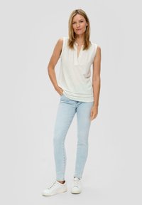 White, sleeveless top with a V-neck and decorative stitching at the hem, paired with light blue jeans and white sneakers.